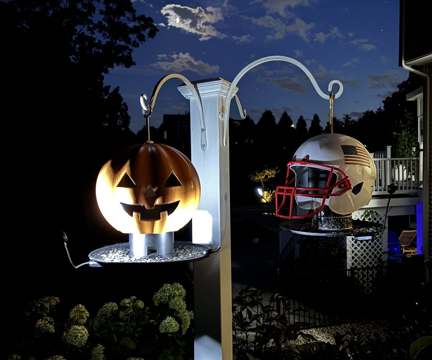 A jack-o-lantern ScoreShell and an American flag football helmet inspired ScoreShell hanging on one pole after dark, both glowing from the light built into their bird feeder bases